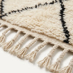 Close-up of a cream-colored textured rug with black geometric border lines and knotted fringe tassels along one edge.