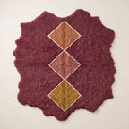 Burgundy Beni Ourain rug with stacked diamond motifs in camel, dusty rose, and gold with cream outlines on a plain background, irregular wavy edges