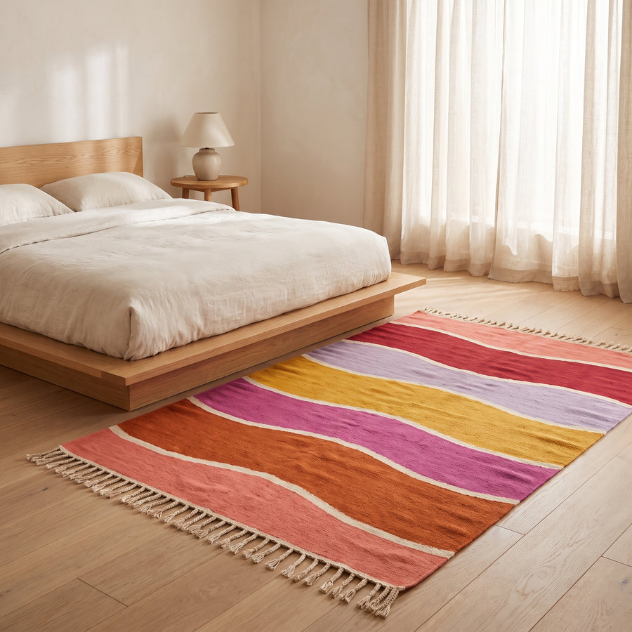 Tamuli rug placed on wooden floor beside a bed with cream linen, showing colour-block stripes in bedroom setting