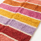 Close-up angled view of Tamuli rug showing soft pile texture and colour transitions between coral, orange, pink, yellow, purple and red stripes