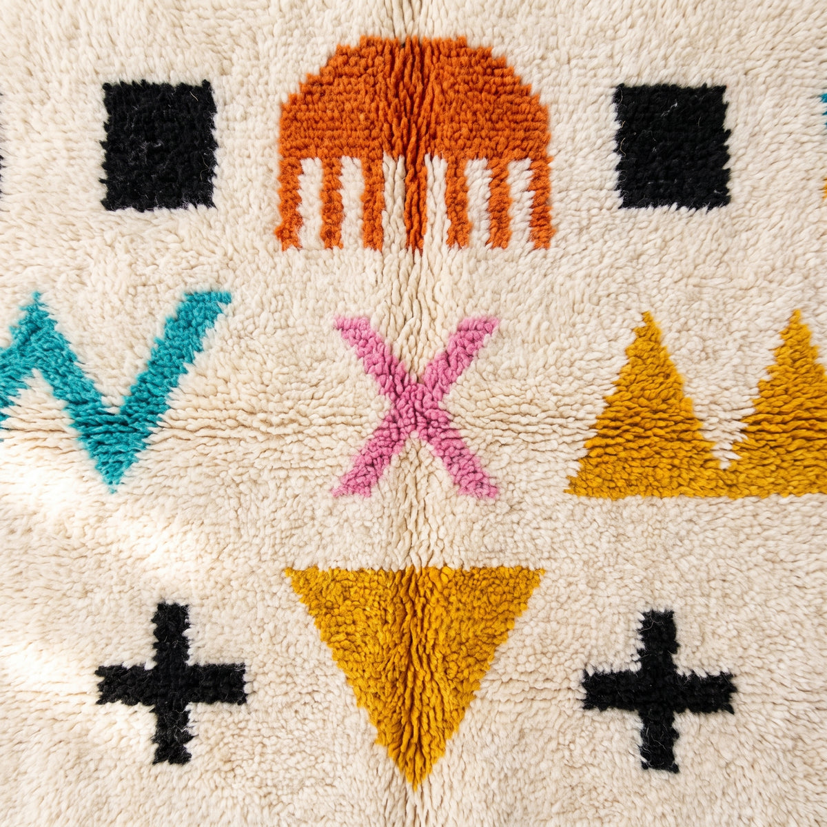 Textured close-up of expressive hand-drawn symbols including black plus signs, magenta X, teal lightning bolts, gold triangles and orange combs in handwoven pile