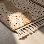 Corner detail of Beni Ourain rug showing cream wool fringe and black diamond motifs at edge