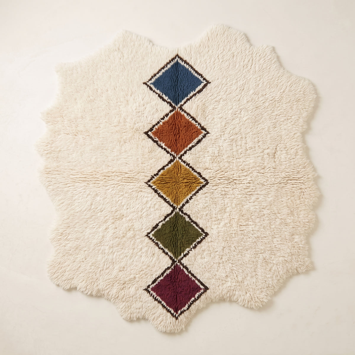 Flat lay of Tafrara Beni Ourain rug with irregular wavy edges, undyed ivory ground and vertical chain of five stacked diamond lozenges in navy, terracotta, gold, olive and burgundy with dark brown and cream outlines