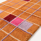 Close-up angled view of the dense, finely knotted plush pile showing orange, pink, red and purple blocks with crisp white lines and periwinkle grid lines