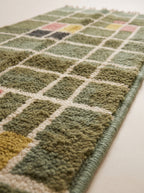 Close-up of the plush hand-knotted pile showing sage green, olive, and cream grid check blocks with pink and mustard accents