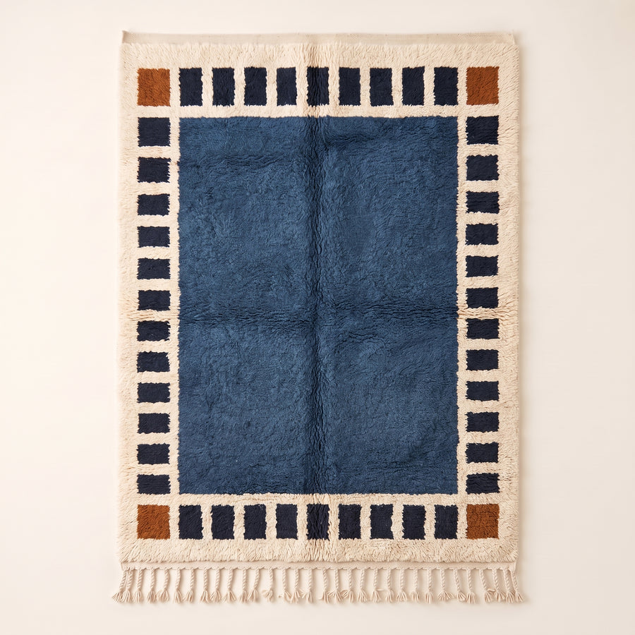 Slate blue Mrirt rug Talwit with navy and rust checkered border and solid center field, flat lay on plain background
