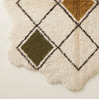 Edge and fringe detail of irregular wavy-edged Beni Ourain rug showing olive and gold squares with cream fringe